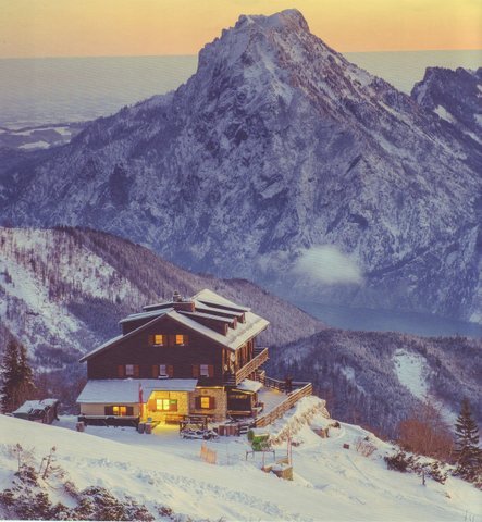 Kranabethhütte glowing at dusk in deep snow, with the mountain rising behind it